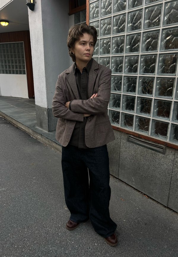 Persona que lleva una chaqueta marrón y una camisa negra está de pie con los brazos cruzados en una acera junto a una pared de bloques de vidrio al anochecer.