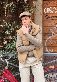 Tan puffer-liivi beigen värisen villapaidan päällä, yhdistetty beigeihin housuihin. Harmaa lippalakki päällä, ja ranteessa näkyy hopeinen rannekoru. Lähellä punainen polkupyörä.