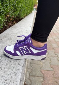 Purple and white New Balance trainers made from suede and leather, showcasing a prominent 'N' logo and textured laces, placed on a kerb.
