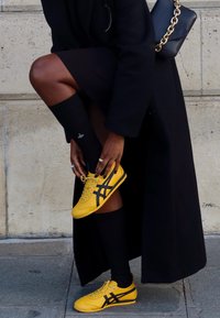 Gelbe Sportschuhe mit schwarzen Akzenten, kombiniert mit schwarzen knielangen Socken, vor einem neutralen Steinhintergrund.