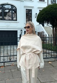 Kremasta teksturirana bluza s remenom i širokim rukavima, uparena sa sjajnim, svijetlim hlačama. Crne sunčane naočale upotpunjuju izgled na pozadini bijele zgrade.