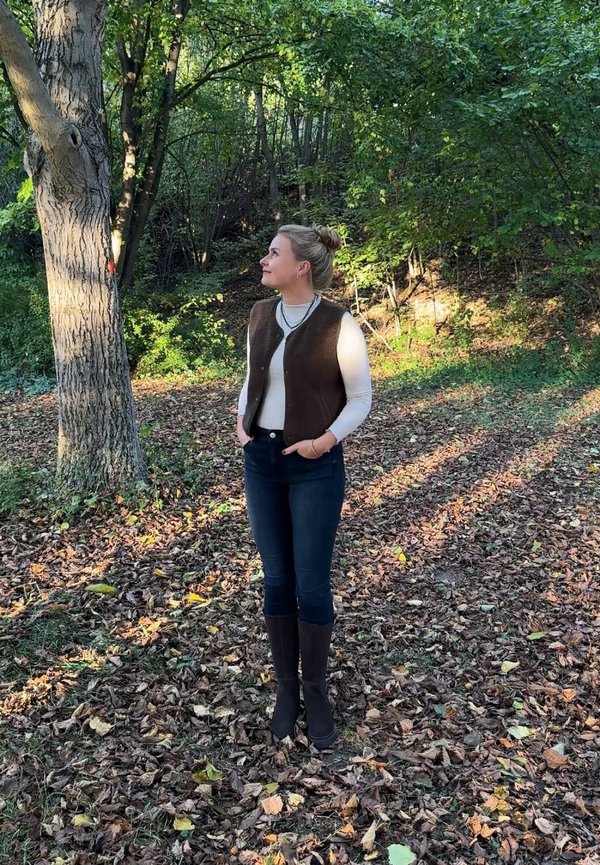 Une femme vêtue d’un gilet brun et de bottes hautes se tient parmi les feuilles mortes dans un sous-bois ensoleillé et regarde un arbre en levant les yeux.