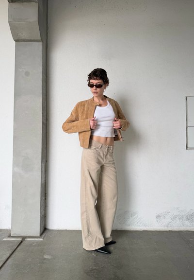 Persona con chaqueta de ante marrón, top corto blanco, pantalones anchos beige, zapatos negros puntiagudos y gafas de sol oscuras, de pie contra una pared blanca.
