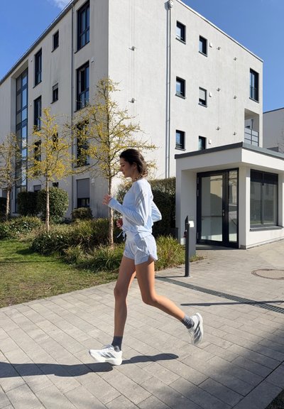 Joven mujer corriendo por un camino pavimentado junto a edificios residenciales modernos con árboles y arbustos en un día soleado.