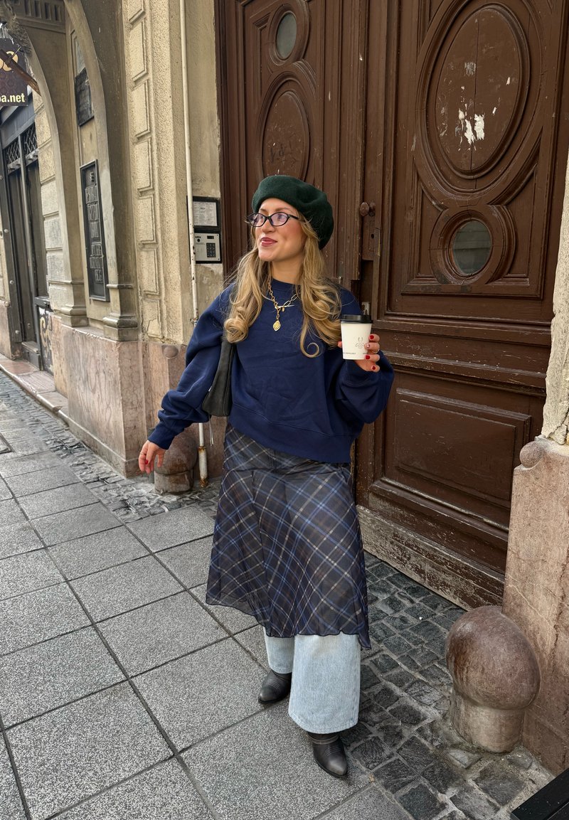 Dunkelblauer oversize Sweatshirt, transparente karierte Rock, weite hellblaue Jeans, schwarze Stiefeletten, grünes Barett, hält eine Kaffeetasse. Steinmauer im Hintergrund.