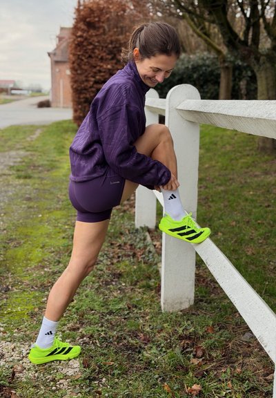 Mujer con ropa deportiva morada estira su pierna sobre una cerca de madera blanca al aire libre, usando zapatillas de correr verde neón y calcetas blancas.