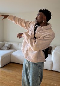 Homme portant une veste beige et un jean effectuant un dab dans un salon avec des canapés blancs et un sol en bois.