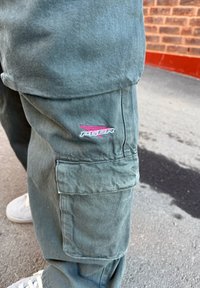 Gröna cargobyxor i slitstarkt material, med sidofickor och en liten rosa och svart logotyp nära fickan. Texturerad yta synlig.