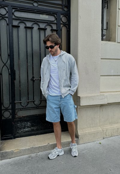 Sudadera gris con cremallera sobre una camiseta blanca, pantalones cortos de mezclilla y zapatillas deportivas grises. Gafas de sol negras y de pie contra una puerta de hierro decorativa.