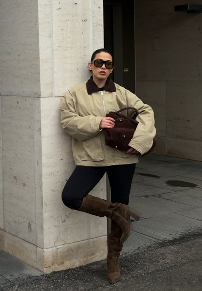 Mujer con chaqueta beige de gran tamaño, leggings negros y botas marrones hasta la rodilla se apoya contra una pared de piedra sosteniendo un bolso marrón, lleva gafas de sol.