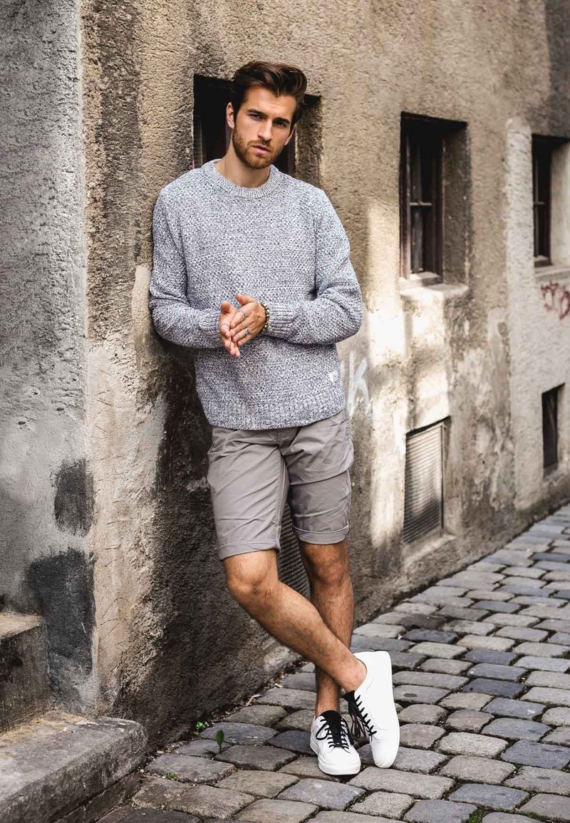 Grijze gebreide trui met textuur, lichtgrijze shorts en witte sneakers met zwarte veters. Staand tegen een gemêleerde muur in een straat met kasseien.