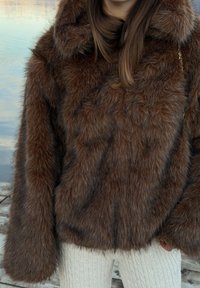 Manteau en fausse fourrure marron à manches longues porté sur un pantalon crème en maille torsadée, sur un fond flou d'eau en extérieur.