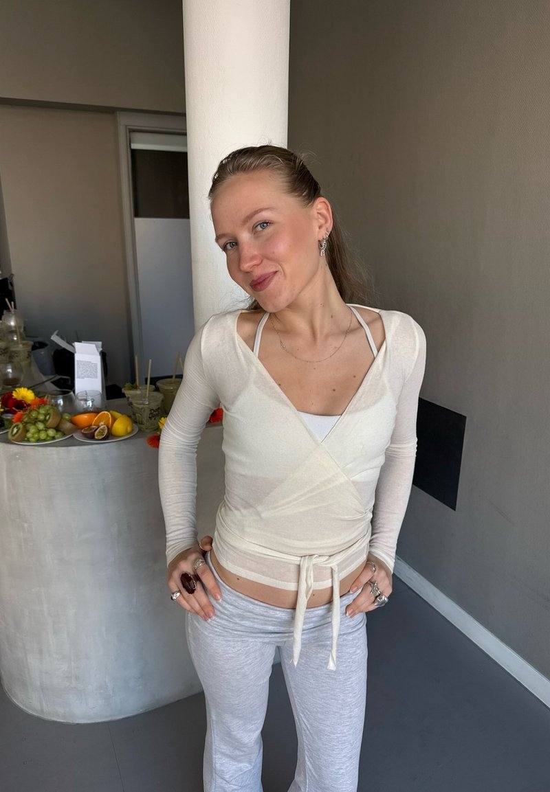 Jeune femme en haut cache-cœur léger et pantalon de survêtement posant avec un sourire près d'une table avec des fruits et des boissons dans une pièce moderne aux tons neutres.