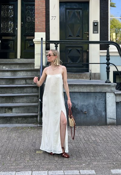 Vestido blanco largo hasta el suelo con patrones florales texturizados. Presenta una abertura lateral, tirantes finos y se complementa con sandalias de tiras marrones y un bolso de mimbre.