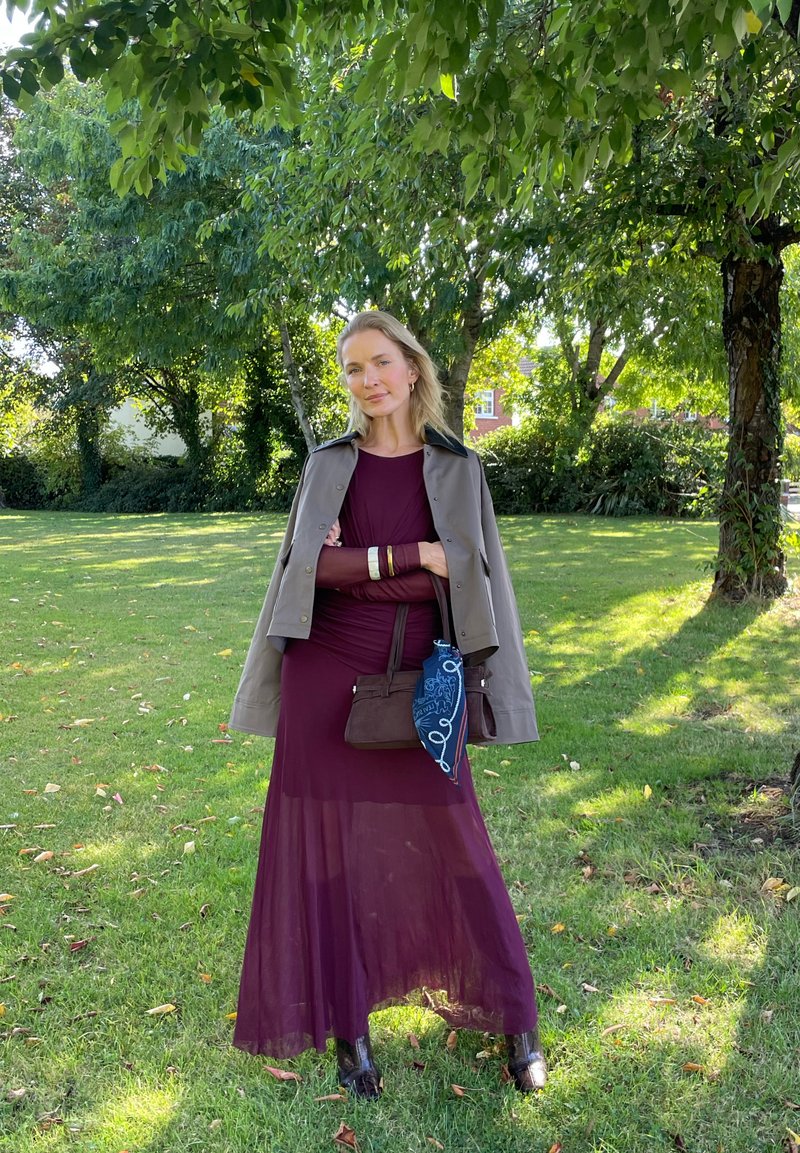 Donna in un lungo abito bordeaux e giacca grigia sta sull'erba sotto alberi foliati, tenendo una borsa marrone con una sciarpa blu.