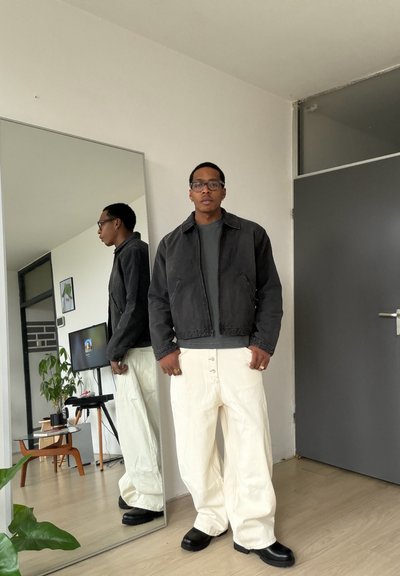 Hombre con chaqueta negra, camisa gris, pantalones blancos y zapatos negros de pie junto a un espejo de cuerpo entero en una habitación moderna.