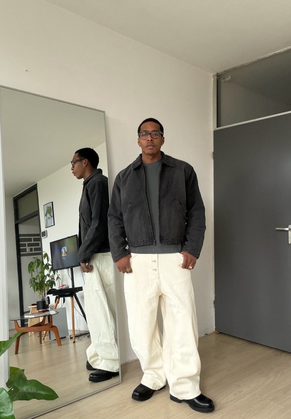 Férfi fekete dzsekiben, szürke ingben, fehér nadrágban és fekete cipőben, aki egy tükör mellett áll egy modern szobában.