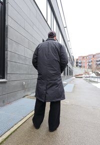 Άτομο που φορά μακρύ σκούρο παλτό και φαρδιά μαύρη παντελόνα, περπατά σε βρεγμένο πεζοδρόμιο δίπλα σε μοντέρνα γκρι κτίριο με γυάλινες παράθυρ ες.