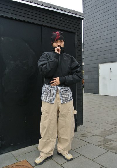 Chaqueta negra con cremallera sobre una camisa a cuadros azul y blanca, combinada con pantalones holgados beige y zapatos beige. Lleva un gorro negro con detalles en rojo.