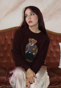 Burgundowy sweter z motywem misia w formalnym stroju, zestawiony z jasnymi satynowymi spodniami. Siedzi na tapicerowanej welurowej sofie.