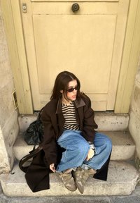 Uma pessoa vestindo um casaco oversized castanho, uma camisa às riscas e calças de ganga largas azuis senta-se em degraus de pedra. Tênis castanhos notáveis e uma bolsa preta estão por perto.