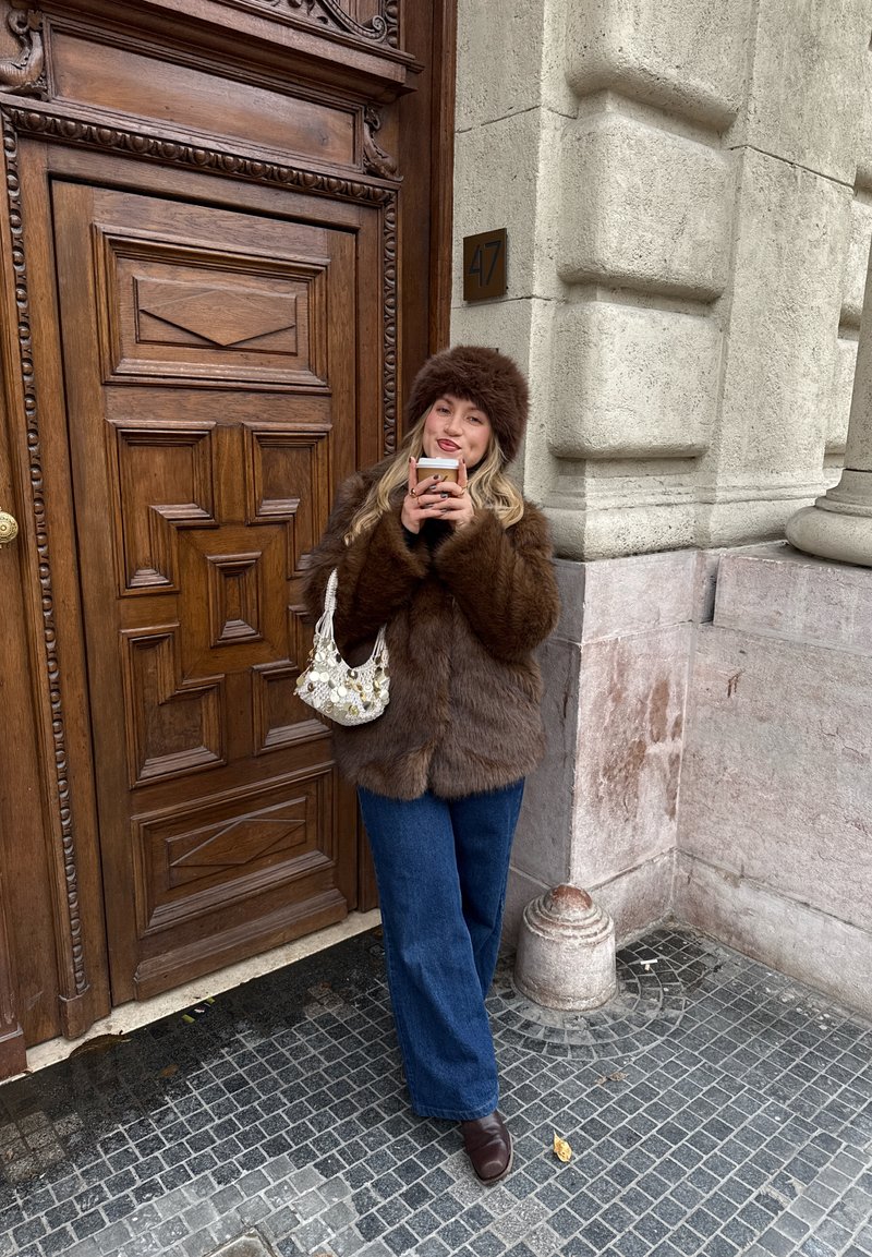 Femme en manteau de fourrure marron et chapeau tenant une tasse de café, debout près d'une porte en bois ornée portant le numéro 47, à l'angle d'un bâtiment en pierre.