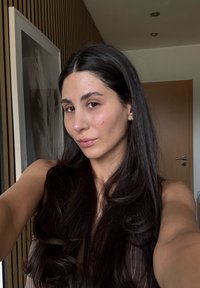Ritratto di una persona con lunghi capelli neri e trucco naturale, che indossa una canotta, posata davanti a una parete e una porta in legno.