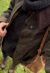 Bruine geparfumeerde jas met een geruite voering, zwarte pluizige trui, corduroy kraag, bruine leren tas en donkere jeans met bruine laarzen.