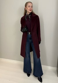 Manteau en laine bordeaux avec revers à encoches, associé à un pull sombre, un jean taille haute à jambes larges, ainsi qu'à des gants et chaussures noirs.