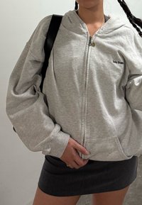 Persoană îmbrăcată într-o hanorac zip-up gri deschis cu textul "lets fran.", geantă de umăr neagră, fustă închisă la culoare și un colier, stând în interior.