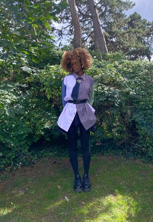 Camisa de gran tamaño con múltiples patrones, rayas y colores sólidos, ceñida a la cintura, combinada con una falda negra y botas negras.