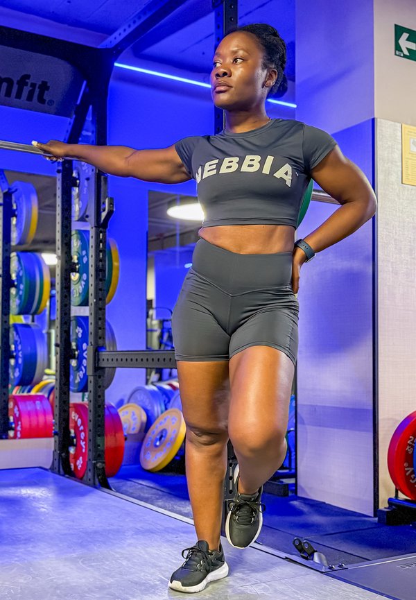 Woman in black athletic wear stretching arm on barbell rack in a gym with colorful weight plates and blue lighting.