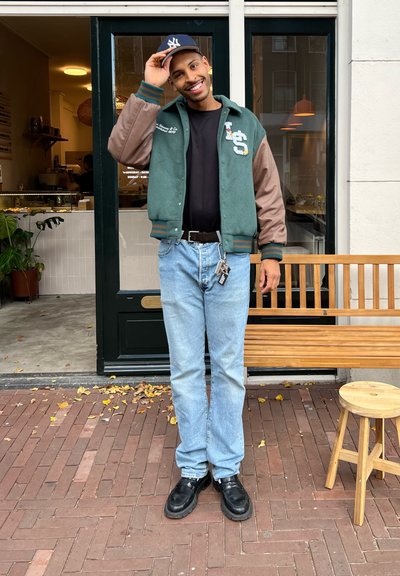 Chaqueta varsity verde y marrón sobre una camiseta negra, jean claros azules y zapatos negros. Entorno al aire libre con un banco de madera y plantas.