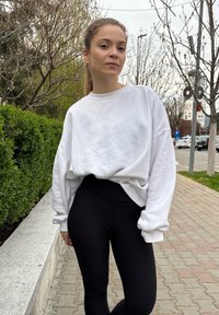 White oversized sweatshirt with dropped shoulders, teamed with black high-waisted leggings. The background includes hedges and pavement tiles.