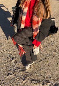 Personne portant un pantalon à rayures, un pull rouge et une écharpe à carreaux ajustant des baskets blanches en étant assise sur une surface de gravier en plein air.