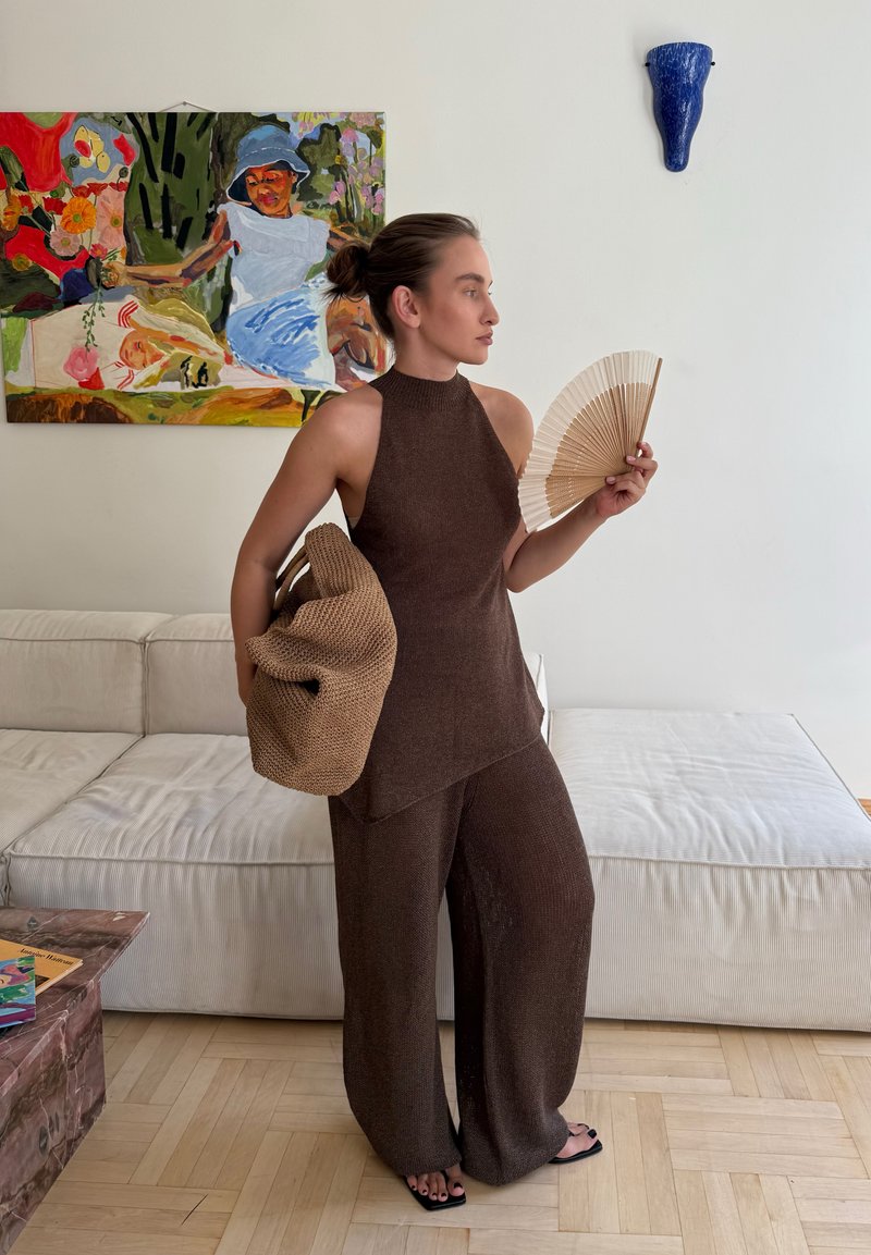 Femme en haut marron sans manches et pantalon tient un sac tissé et un éventail à la main, debout dans un salon avec un canapé et un tableau coloré.