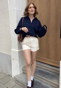 Navy zip-up sweatshirt, creamfärgade shorts, brun bälte och väska, vita ankelsockar och vinröda loafers. Utomhusmiljö med bakgrund av en träddörr.