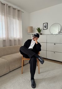 Mann mit schwarzer Mütze, Brille und Kleidung sitzt im Schneidersitz auf einem Holzstuhl in einem minimalistischen Wohnzimmer mit beigem Sofa und weißem Schrank.
