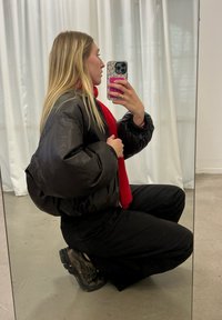 Casaco de puffer preto com um design curto, combinado com um cachecol vermelho brilhante. Calças e sapatos pretos com sola texturizada. Reflexo no espelho visível.