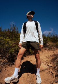 Conjunto atlético com uma camisa branca de mangas longas, calções verdes escuros, meias brancas e ténis brancos robustos. Ambiente exterior com vegetação.