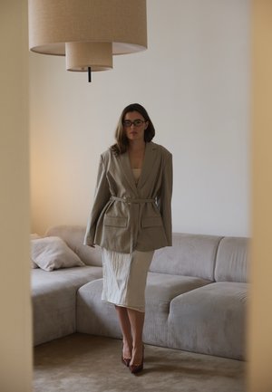 Woman wearing oversized beige blazer with belt, fitted light skirt, and glasses stands in minimalist living room with beige sofa and pendant lamp.