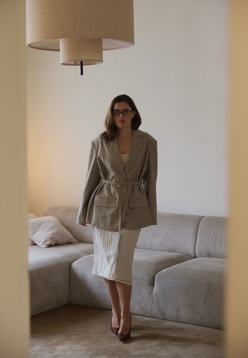 Frau in übergroßem beigen Blazer mit Gürtel, passendem hellen Rock und Brille steht in einem minimalistischen Wohnzimmer mit beigem Sofa und Pendelleuchte.