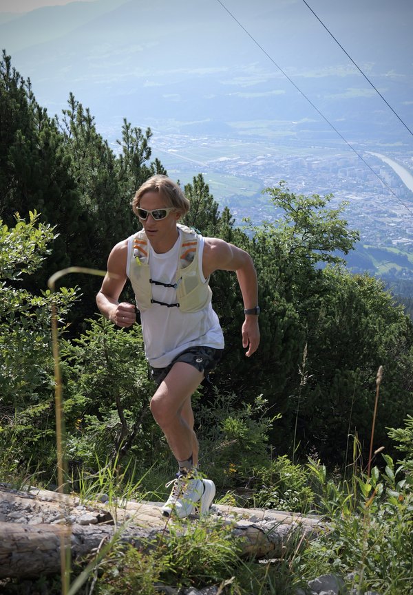 Man wearing sunglasses and running gear running uphill on a forest trail with mountains and a city visible in the background.