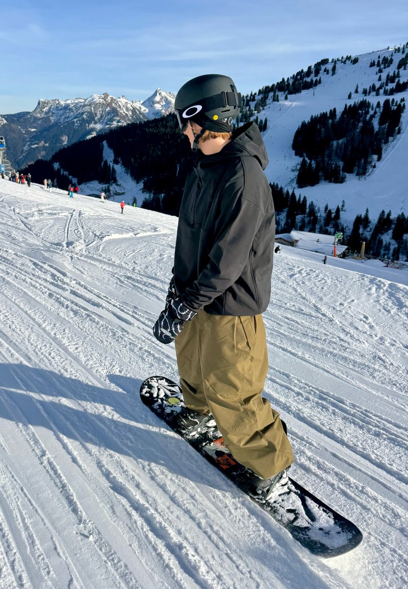 Snowboardeur sur une pente enneigée, portant une veste noire à capuche, un pantalon beige, des gants noirs et un casque noir avec un accent blanc.