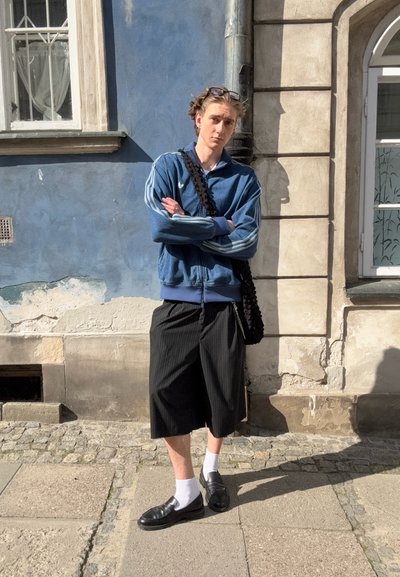 Chaqueta azul con cremallera y rayas blancas, pantalones cortos de rayas finas negras, calcetines blancos, zapatos negros y una bolsa negra texturizada. De pie contra una pared azul.