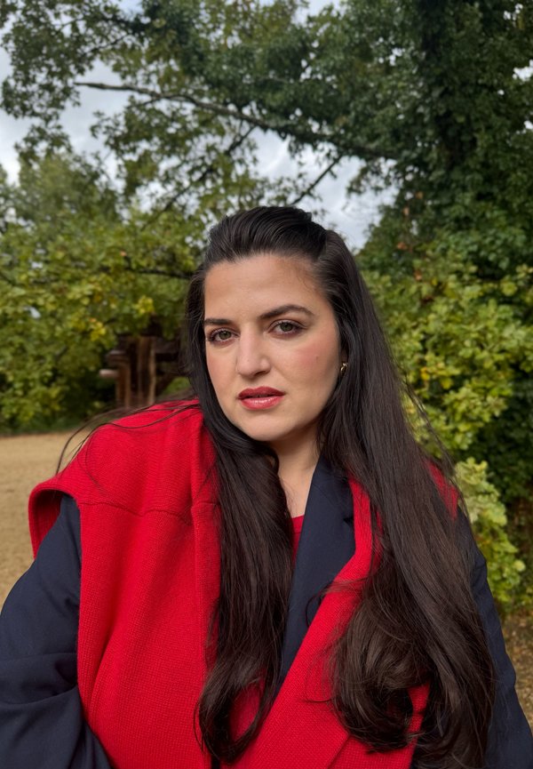 Wrap in maglia rossa sopra una giacca blu, con lunghi capelli scuri. Sullo sfondo si vedono fogliame verde sotto un cielo nuvoloso, creando un ambiente naturale.