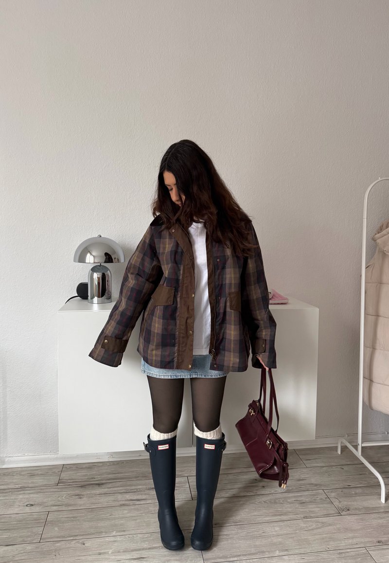 Donna con giacca a quadri, gonna di jeans, calze nere e stivali da pioggia neri che tiene una borsa color bordeaux in casa vicino a mobili bianchi.