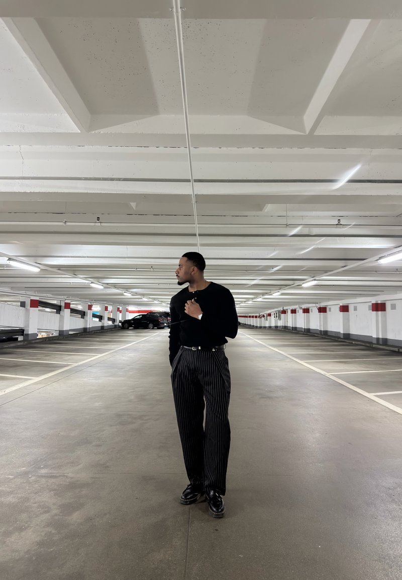 Homme en pull noir et pantalon à fines rayures debout dans un parking à plusieurs niveaux vide, regardant vers sa gauche.