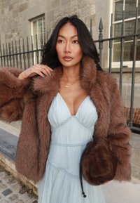 Rudas dirbtinis kailinis paltas su plačiu apykakle, dėvimas virš šviesiai mėlynos v formos suknelės su tekstūruotu audiniu ir nėrinių akcentais. Laikoma ruda rankinė.