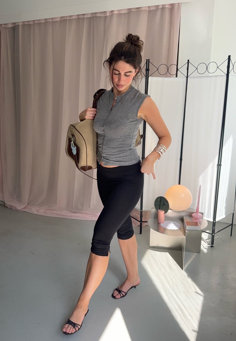 Junge Frau in grauem ärmellosen Top und schwarzen Capri-Leggings hält eine beige Tasche und posiert drinnen neben einem dekorativen Raumteiler und einem Tisch mit Lampe.